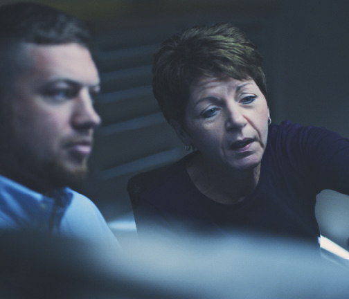 Two people, a man and a woman, are sitting closely together, looking intently at something off-camera. The woman appears to be explaining or pointing something out to the man. The setting seems to be an office or workspace.